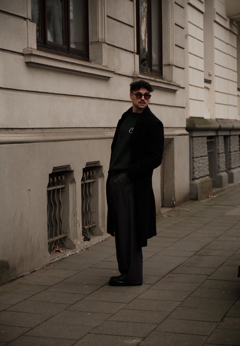 Cappotto nero, maglione verde, pantaloni grigi e scarpe scure. Capelli ricci, occhiali da sole, in piedi vicino a un edificio con dettagli in pietra.