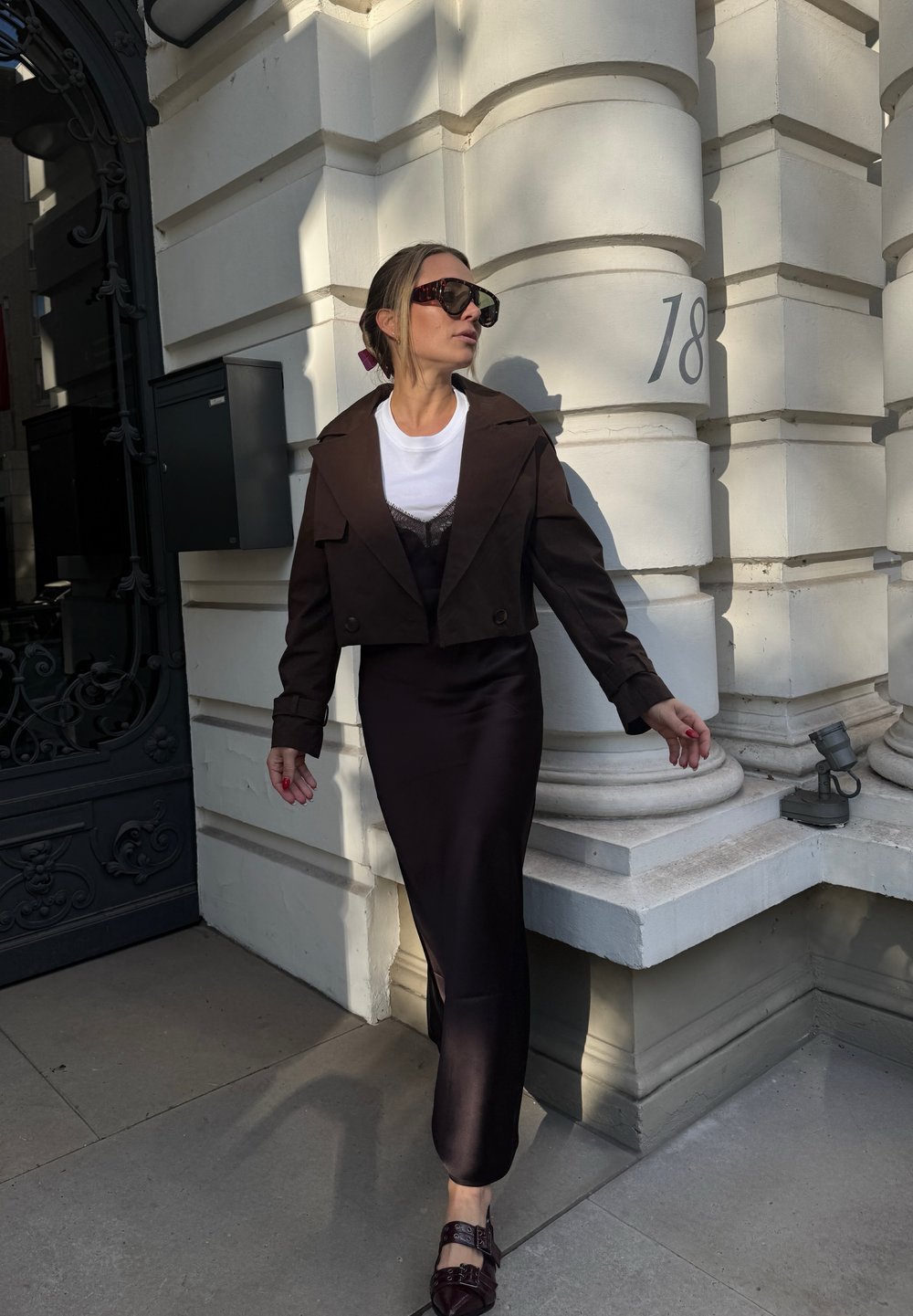 Giacca corta marrone su una t-shirt bianca e una gonna nera in satin con uno spacco laterale. Gli occhiali da sole neri e le scarpe bordeaux completano il look.