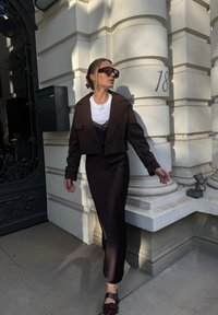 Giacca corta marrone su una t-shirt bianca e una gonna nera in satin con uno spacco laterale. Gli occhiali da sole neri e le scarpe bordeaux completano il look.