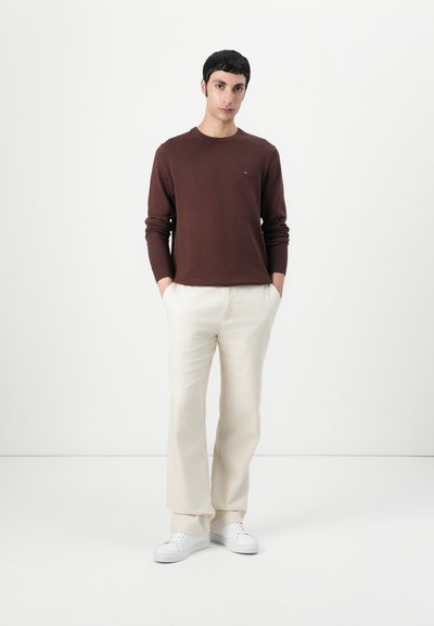 Suéter de punto marrón, pantalones crema, zapatillas blancas. Modelo de pie con las manos en los bolsillos contra un fondo blanco liso. Diseño simple, corte casual.