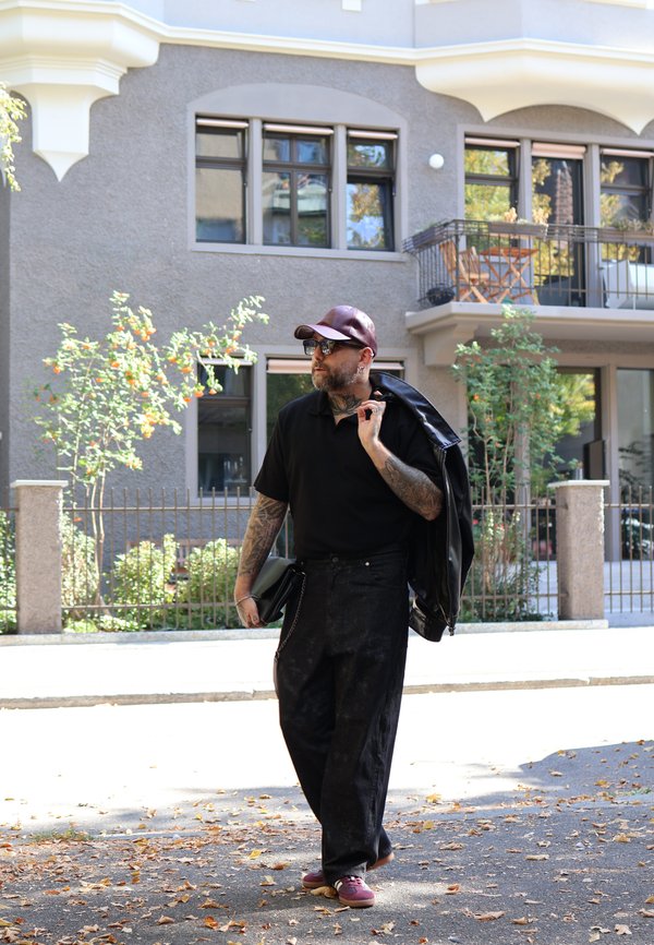 Hombre con tatuajes y gafas de sol lleva una chaqueta negra sobre el hombro y una bolsa, caminando por una acera cubierta de hojas cerca de un apartamento.