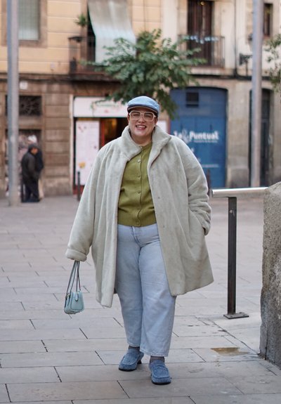 Persona sonriente caminando por una calle de la ciudad, que lleva un abrigo ligero de piel, suéter verde, jeans azul claro, zapatos planos, gorra y un pequeño bolso.