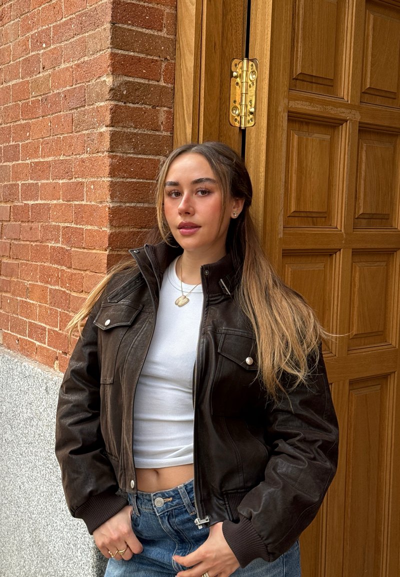 Jeune femme aux longs cheveux debout contre un mur de briques et une porte en bois, portant une veste sombre, un crop top blanc et un jean bleu.