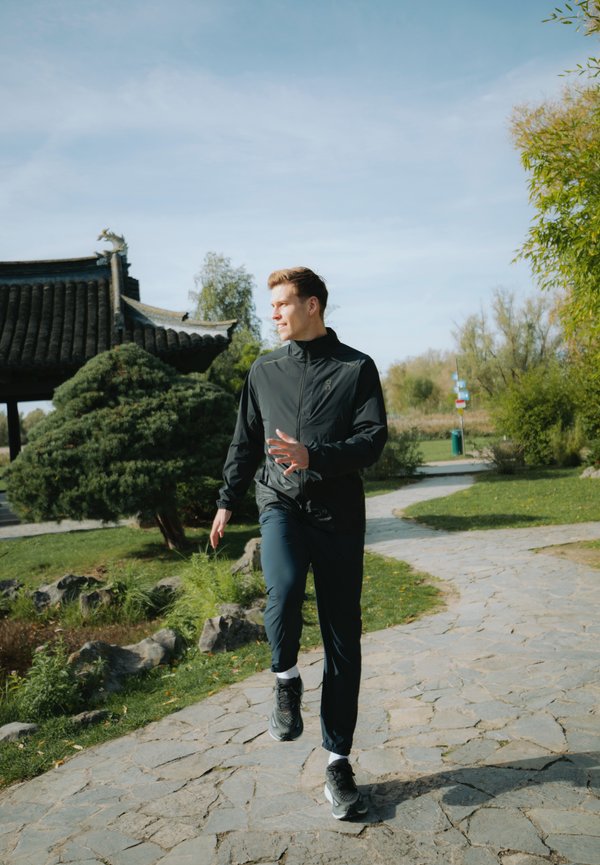 Chaqueta deportiva negra con cremallera, combinada con pantalones azul oscuro. Persona que lleva zapatillas negras, caminando por un sendero de piedra en un parque.
