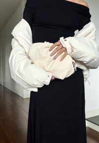 Crna haljina s otpadom na ramenima u kombinaciji s bujnim bijelim puffer jaknom. Drži meku clutch torbicu u krem boji.