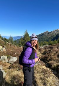 Una donna con giacca viola e berretto grigio, indossando occhiali da sole, fa escursionismo su un sentiero montano roccioso sotto un cielo blu chiaro.