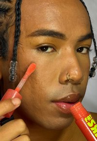 Personne avec des cheveux tressés applique un brillant à lèvres orange vif sur la joue, tenant un tube de brillant à lèvres ouvert étiqueté "Fat Smo."