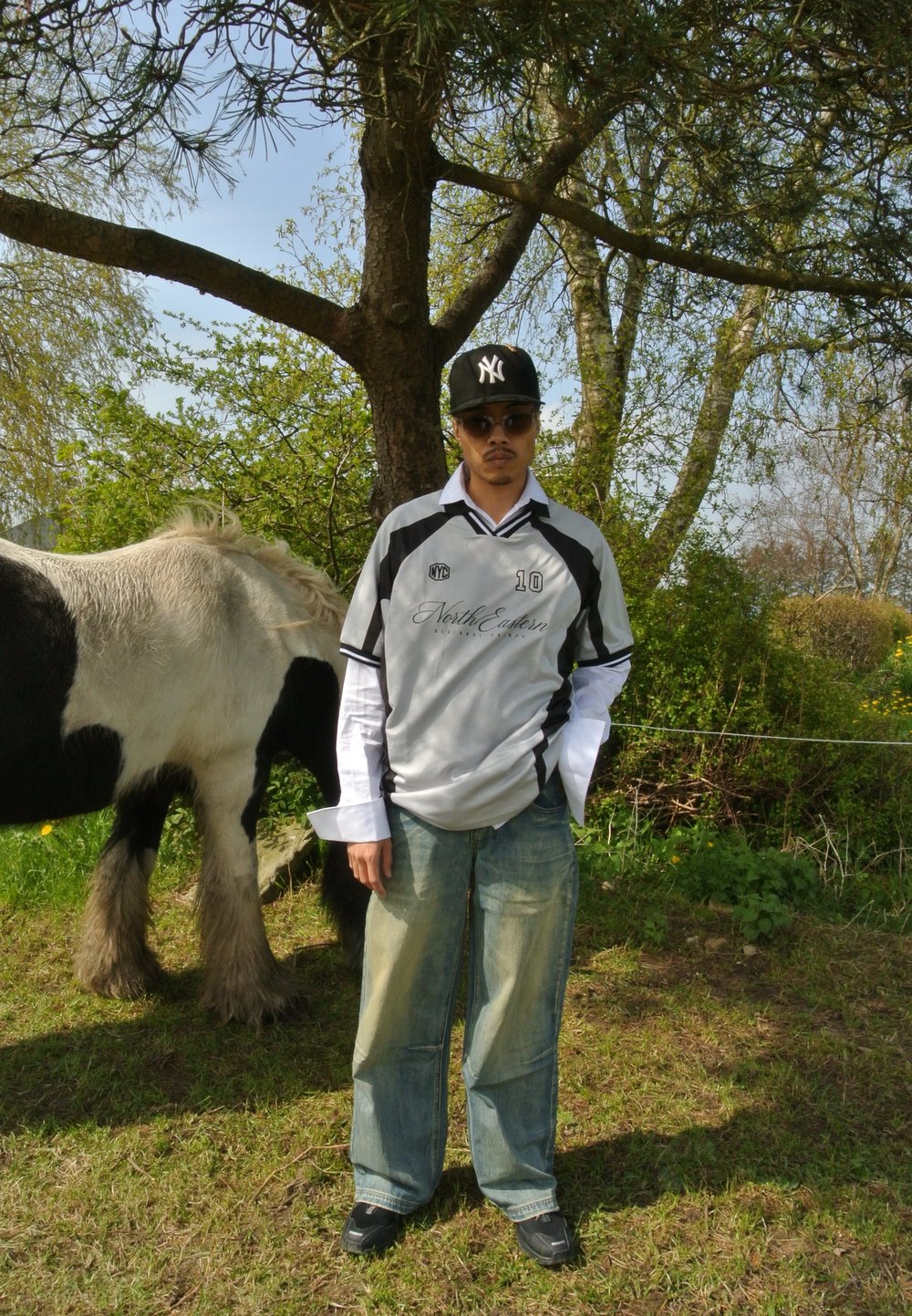 Graues und schwarzes Sporttrikot mit der Nummer 10, lockere blaue Jeans, Sonnenbrille und eine schwarze Mütze, stehend neben einem schwarz-weißen Pony in einem grasbewachsenen Bereich.