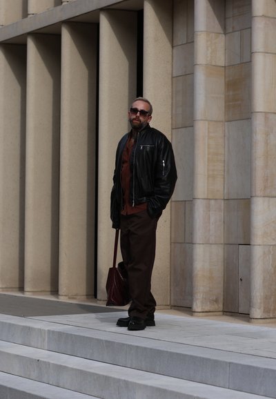 Hombre con gafas de sol, chaqueta de cuero negra y pantalones marrones sosteniendo una bolsa, de pie junto a grandes columnas de piedra beige al aire libre.