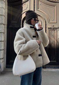 Casaco de fleece bege com botões pretos, segurando uma chávena branca. Combinado com uma mala hobo branca e jeans de ganga azuis. Gorro escuro e óculos de sol.
