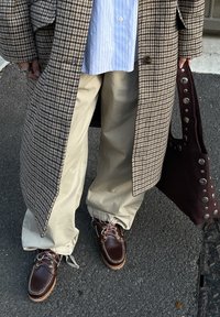 Personne portant un pantalon large beige, des chaussures en cuir marron, une chemise bleue à rayures et un manteau à motif pied-de-poule, tenant un sac clouté marron foncé.