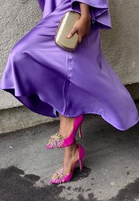 Scarpe con tacco alto rosa con dettagli decorativi di perline e punta aperta, abbinate a un abito viola fluente e una borsa a tracolla dorata.