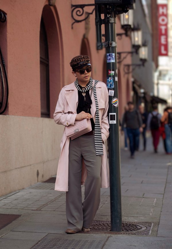 Lichtroze trenchcoat, zwarte blouse, grijze wijde broek, zwart-wit gestreepte sjaal en een lichtroze handtas. Leeuprint hoed en donkere zonnebril.