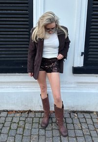 Blazer in velluto a coste marrone, camicia bianca aderente, pantaloncini neri con paillettes e stivali marroni al ginocchio con fibbie. Pavimentazione in ciottoli.