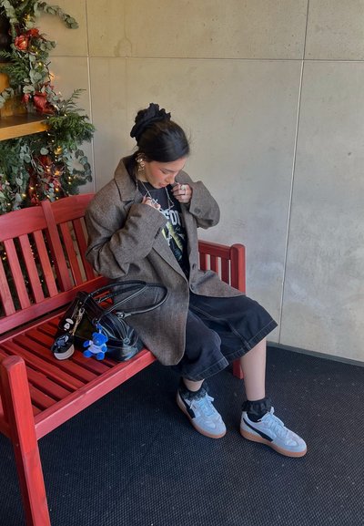 Abrigo grande marrón sobre una camiseta gráfica negra, falda de denim negra, zapatillas gris con suelas de goma, sosteniendo un bolso negro en un banco rojo.