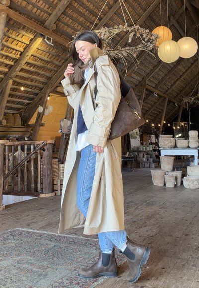 Mujer con gabardina beige, vaqueros y botas marrones está dentro de un loft rústico de madera con cerámicas y faroles colgantes.