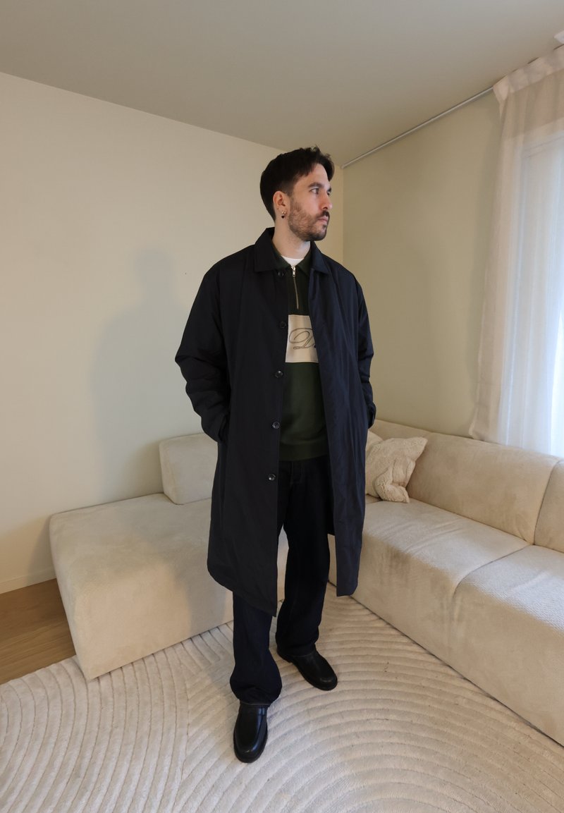 Homme aux cheveux courts et foncés et à la barbe, portant un long manteau noir et un pantalon sombre, debout dans un salon beige avec un canapé et des rideaux transparents.