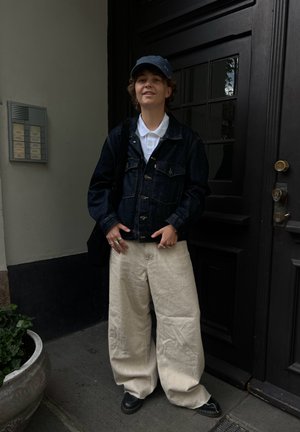 Chaqueta de mezclilla sobre un polo blanco, pantalones de pierna ancha beige, zapatos negros. Cabello corto y rizado, usando una gorra azul. De pie junto a una puerta negra.