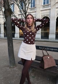 Jeune femme portant un haut à pois marron, une jupe beige et des collants foncés pose avec les mains derrière la tête près d’un arbre et d’un banc avec un sac à main marron.