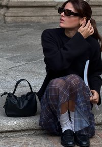 Zwarte oversized trui, doorzichtige geruite rok met blauwe en bruine tinten, witte sokken, zwarte loafers en een kleine zwarte handtas.