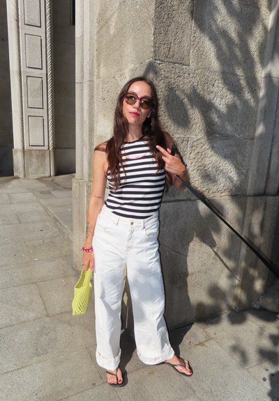 Top sin mangas a rayas, pantalones anchos blancos, bolso amarillo, chanclas. Cabello largo, gafas de sol, de pie contra una pared de piedra texturizada.