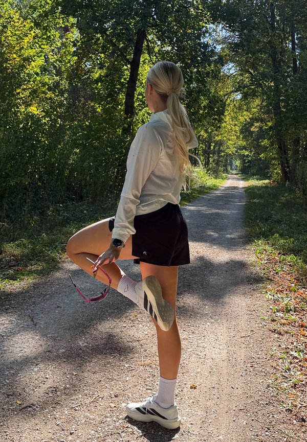 Жена с руса опашка, носеща бяло яке и черни шорти, разтягаща крака на слънчев горски път, държейки слънчеви очила.