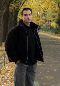 Fekete puffertöltött dzseki magas gallérral, cipzáras záródással, sötét pulóver fölött viselve. Szürke, vékony csíkos nadrág; a háttérben őszi lombok láthatók.