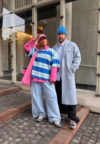 Dos personas están de pie en una acera de piedra junto a un edificio; una viste ropa colorida y de gran tamaño con una gorra rosa, y la otra lleva un gorro azul y un abrigo gris.