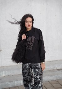 Melnas garām piedurknēm krekliņš ar grafisko apdruku, saskaņots ar rakstainām pelēkām platām biksēm. Rokā tur mīkstu melnu somiņu. Betons fons.