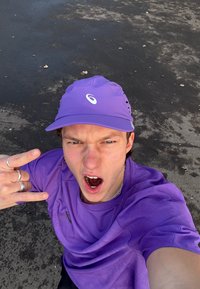 Casquette de sport violette et chemise texturée assortie. La casquette arbore un logo ; la personne fait un geste de la main devant un fond en asphalte.