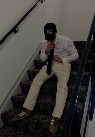 Camisa blanca, corbata negra y pantalones crema en una persona sentada en escaleras con un patrón. Gorro verde y zapatillas oliva con rayas blancas.