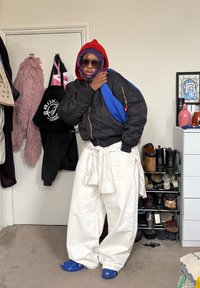 Personne portant une veste bomber noire, un pantalon blanc ample, un couvre-chef rouge et violet, des lunettes de soleil et des chaussures bleues, debout dans une chambre près d'un porte-chaussures et de crochets.