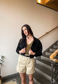 Črna denim jakna z dvema sprednjima žepoma, oblečena na rebrovani olivno zeleni majici, v kombinaciji z bež kratkimi hlačami; gladka tekstura in sproščen kroj.