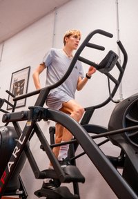 Trainer eliptic negru cu un cadru vertical, pedale mari și monitor. Utilizator îmbrăcat în echipament sportiv gri este în mișcare. Mediu de sală de sport vizibil.