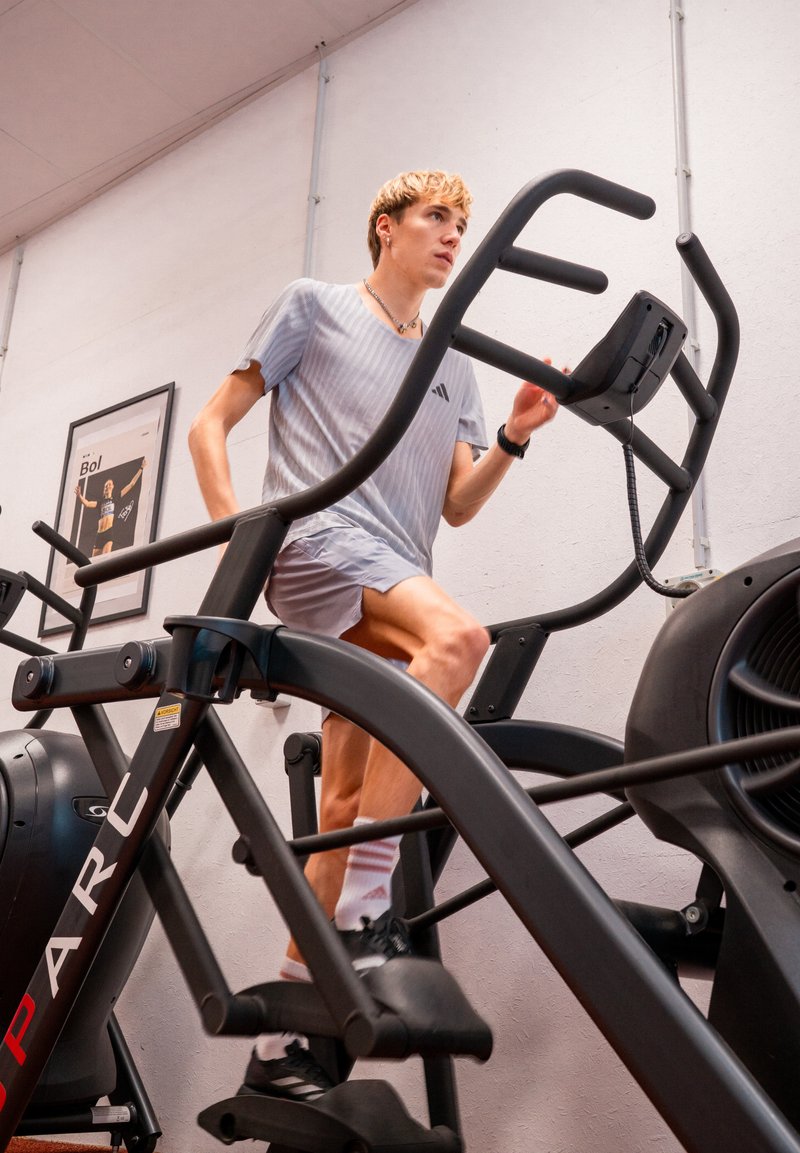 Black elliptical trainer with a vertical frame, large foot pedals, and monitor. User in gray athletic wear is in motion. Gym environment visible.