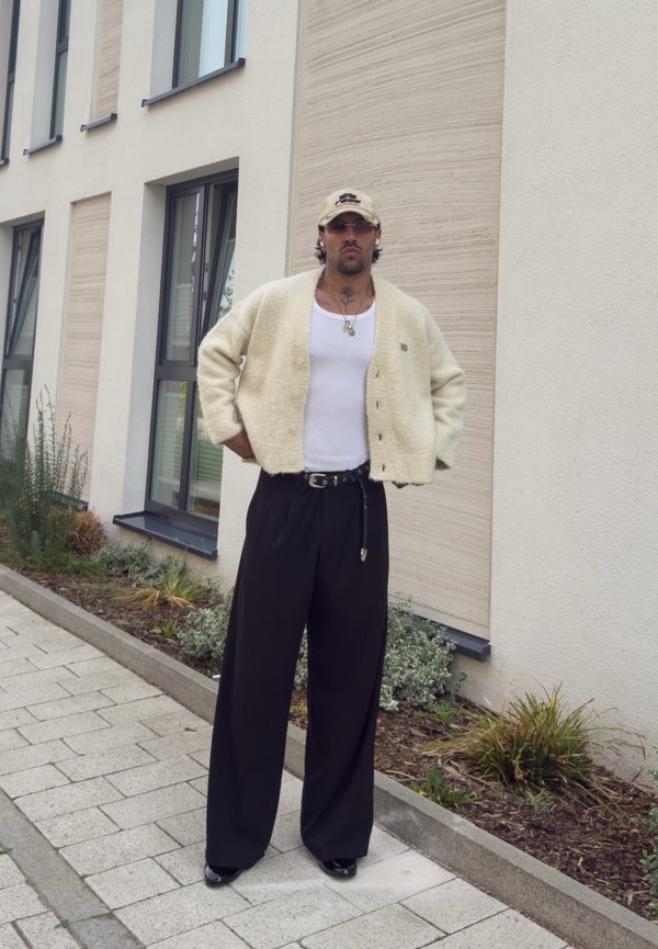 Mann trägt cremefarbenen Cardigan, weißes Tanktop, schwarze weite Hose, beige Kappe und Sonnenbrille, steht auf dem Gehsteig neben modernem Gebäude.