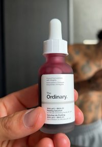 Eine Hand hält die Flasche von The Ordinary AHA 30 % + BHA 2 % Peelinglösung, im Hintergrund ist ein verschwommener, tätowierter Oberkörper zu sehen.