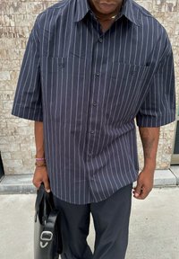 Camicia azzurra scuro button-up con strisce verticali bianche, maniche corte e due tasche sul petto, abbinata a pantaloni neri e una borsa nera.