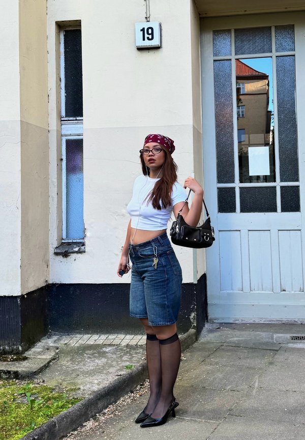 Joven mujer con gafas, bandana roja, top corto blanco, shorts vaqueros, calcetines transparentes hasta la rodilla y tacones, está de pie junto a la entrada del edificio número 19 sosteniendo un bolso negro.
