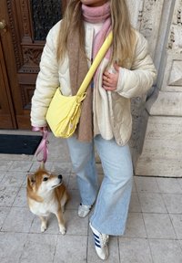 Personne en veste légère matelassée et écharpe multicolore tenant une laisse rose attachée à un petit chien brun et blanc assis sur un sol en pierre.