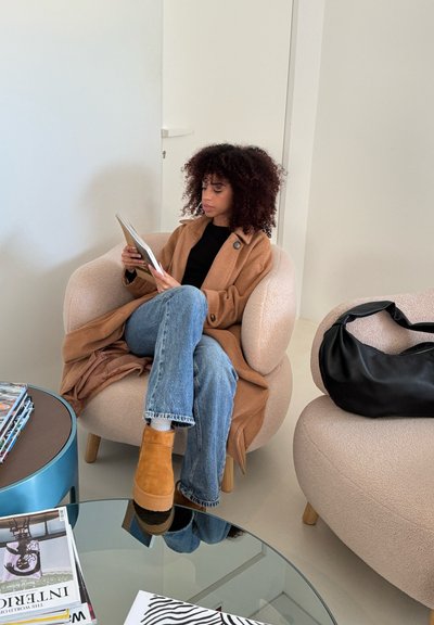Sillón de bouclé beige con forma redondeada. Persona con un abrigo beige y jeans azules, leyendo una revista. Bolso negro a un lado.