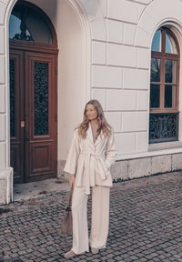 Veste portefeuille couleur crème avec manches larges et pantalon à jambes larges assorti. Texture douce et lisse ; associé à un sac cabas gris. Pavé en pierre de taille.