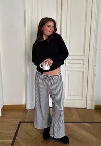 Μαύρο, μακρυμάνικο cropped τοπ, ριγέ παντελόνι σε μαύρο και άσπρο, κρατώντας μια λευκή κούπα. Λείος υφασμάτινος σχεδιασμός, casual στυλ.