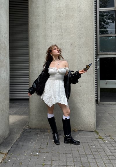 Vestido blanco sin hombros con cuerpo fruncido y dobladillo en capas; chaqueta de piel sintética negra; botas negras hasta la rodilla; sosteniendo gafas de sol; entorno al aire libre.