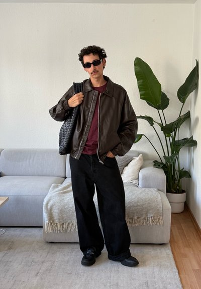 Joven con gafas de sol, vestido con chaqueta de cuero marrón, camisa burdeos, pantalones anchos negros, sosteniendo un bolso negro tejido, de pie en una sala de estar.