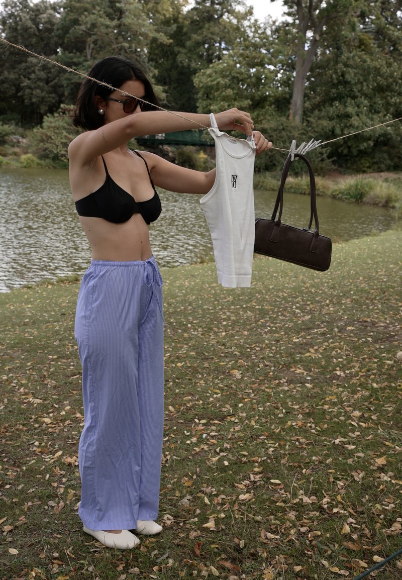 Top bikini nero, top in cotone bianco con logo, pantaloni a righe blu chiaro e una borsa marrone esposti su un filo intrecciato vicino a un lago.
