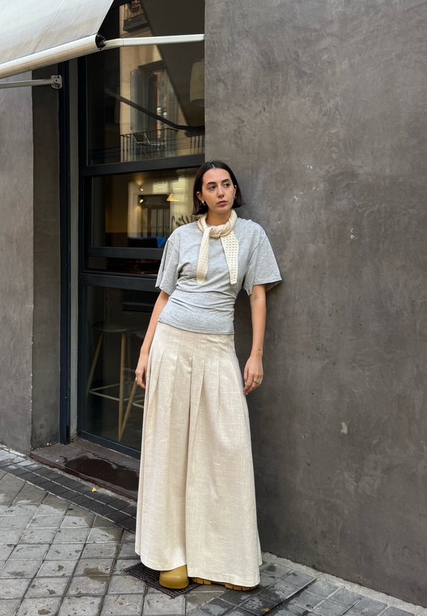 Gray short-sleeve top with a knitted off-white scarf, paired with a long pleated cream skirt and yellow platform shoes, against a concrete wall.