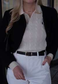 Blazer en laine noire sur un pull crème en maille torsadée, pantalon blanc et ceinture noire avec boucle dorée. Main visible dans la poche, contrastes subtils de textures.