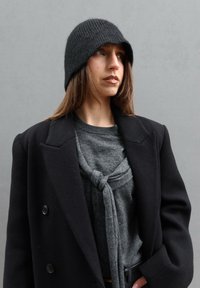 Femme portant un bonnet en maille noir, un manteau sombre surdimensionné et un pull gris noué à l'avant, regardant vers sa droite sur un fond gris uni.