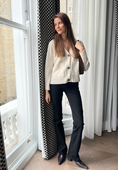 Cardigan crema con cierre de botones y bolsillos frontales, combinado con pantalones negros acampanados y zapatos de punta, de pie cerca de cortinas a cuadros.